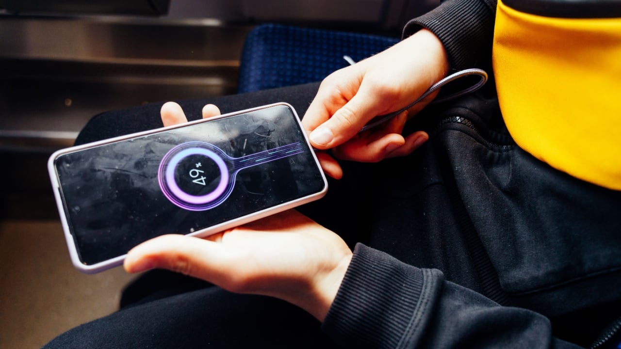 Smartphone charging in teenager's hands, phone shows battery percentage, seated on train in dark clothes and yellow bag, close-up detail of tech usage in public transport