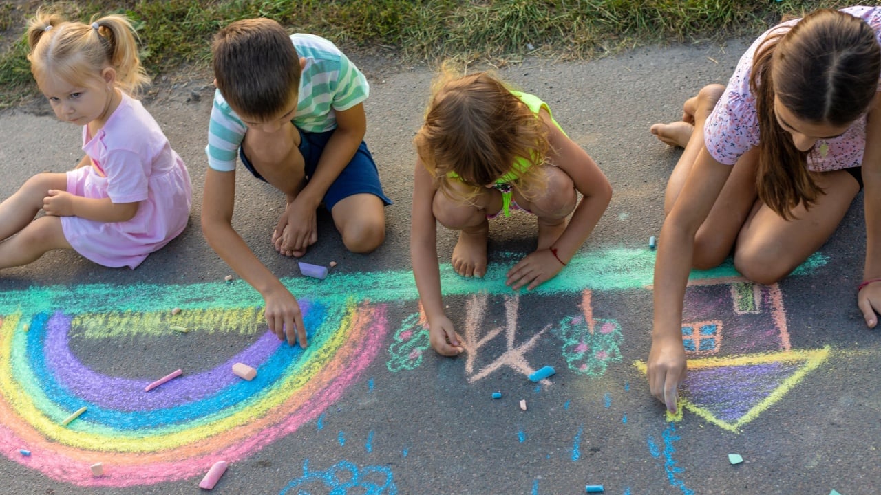 Children draw a picture with chalk on the asphalt. A rainbow, house, sun, clouds are drawn on the asphalt.