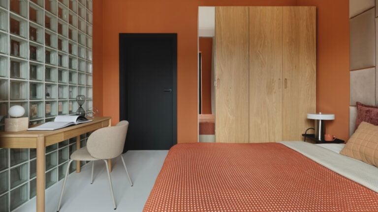 Modern bedroom with orange walls, upholstered geometric headboard, wooden wardrobe, and work desk with glass block wall.
