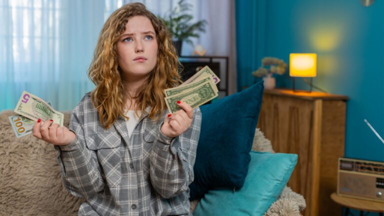 Young woman at home holding cash dollars in hands counting money and thinking about finances with emotion. Caucasian redhead girl on sofa looking satisfied or concerned, personal budget reflection
