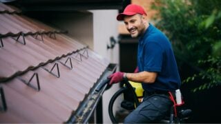 Happy man using leaf blower while cleaning gutters on the roof of a house.