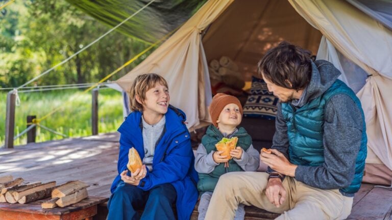 Father with his toddler and teenage son tourists in warm vests and hats eating buns while sitting on glamping steps, enjoying family travel, outdoor adventure, and bonding
