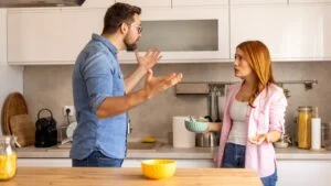 Intense disagreement between a couple in their kitchen. The man raises his hand in a defensive manner. Both display strong emotions, reflecting a challenging moment in their relationship