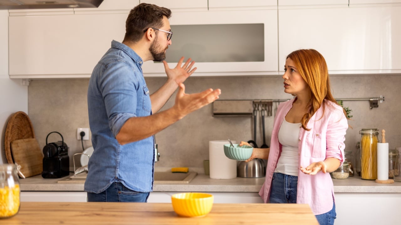 Intense disagreement between a couple in their kitchen. The man raises his hand in a defensive manner. Both display strong emotions, reflecting a challenging moment in their relationship