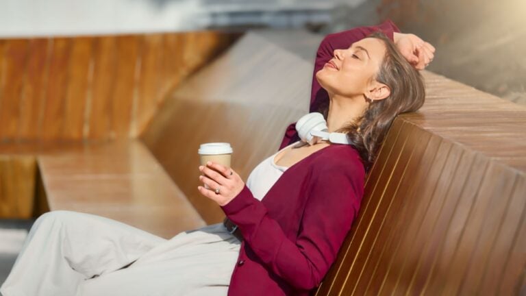 A stylish, relaxed woman leans back on a wooden bench, sipping coffee and soaking in warm sunlight. She wears a burgundy cardigan with a white top and headphones around her neck, radiating calmness.