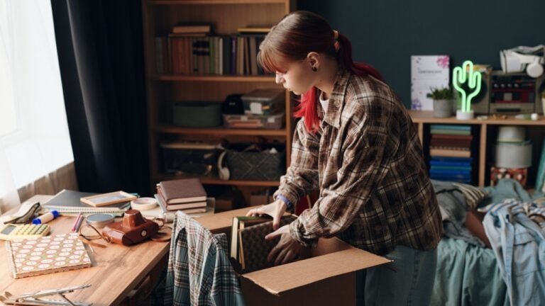 Caucasian teenage girl packing books into cardboard box in bedroom, standing by cluttered desk with various school supplies and personal items, bookshelf in background