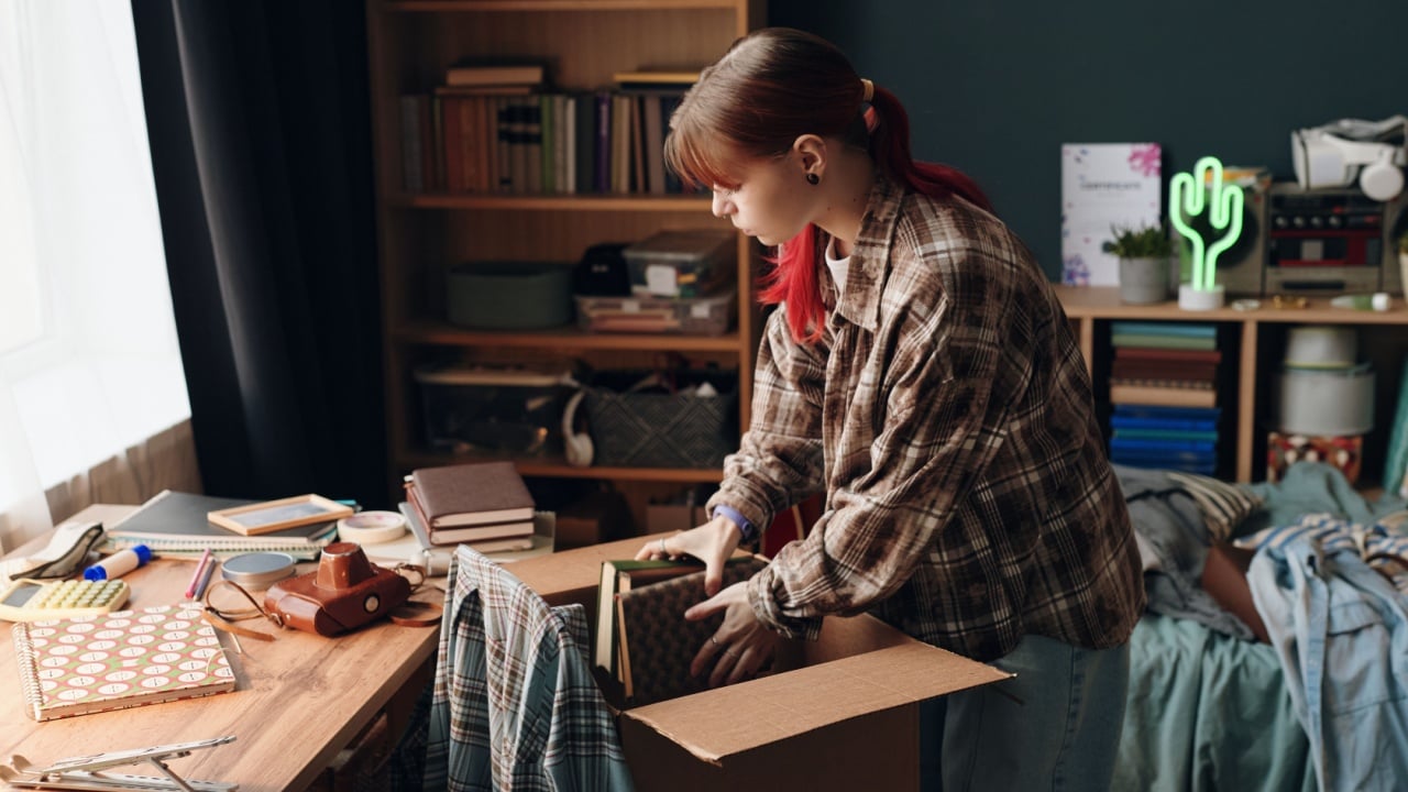 Caucasian teenage girl packing books into cardboard box in bedroom, standing by cluttered desk with various school supplies and personal items, bookshelf in background