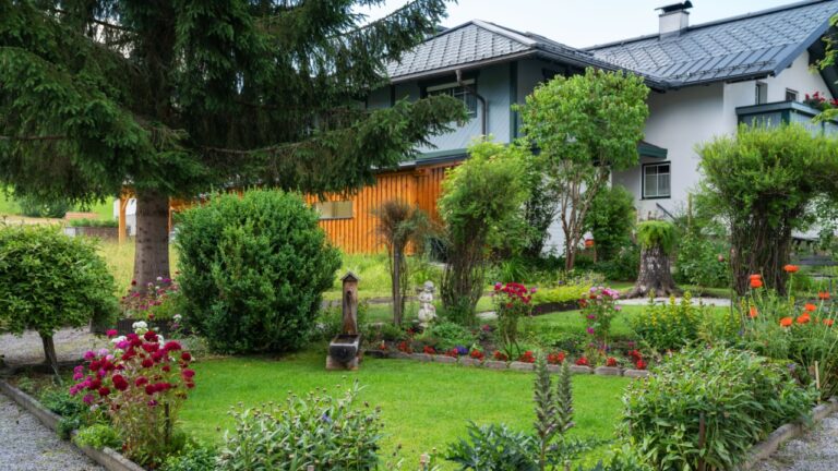 A beautifully kept garden with vibrant flowers, lush green shrubs, and a small wooden water fountain spreads in front of a charming alpine-style house. Decorative details and colorful plants.