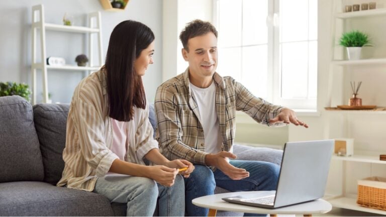 Portrait of a young smiling married couple sitting on sofa at home looking at laptop discussing ideas about interior design. Man and woman planning repair. Renovation and moving concept.