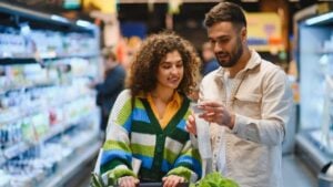 Young couple shopping for groceries in a supermarket, man reading a shopping list while woman pushes the cart filled with produce
