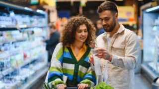 Young couple shopping for groceries in a supermarket, man reading a shopping list while woman pushes the cart filled with produce