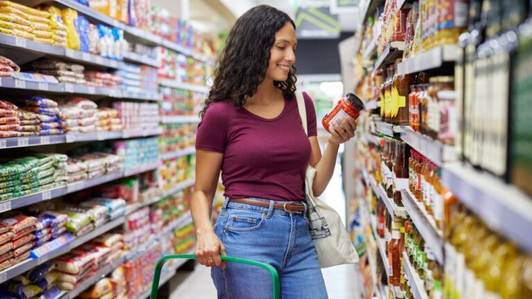 Confident mixed race woman holding jar of tomato soup while standing in grocery store. Young casual woman smiling while looking nutrition label while holding product. Shopper with basket checking ingredients on canned tomato in grocery store.
