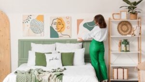 Young woman hanging painting on light wall in bedroom, back view