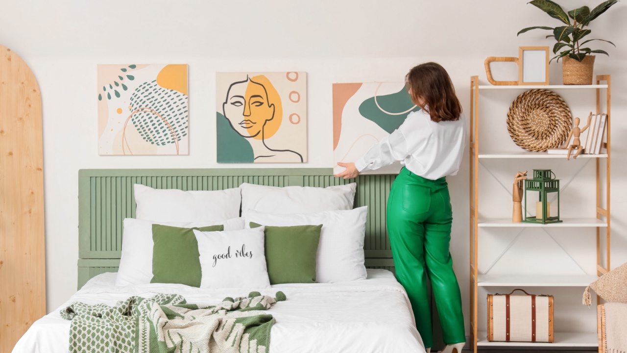 Young woman hanging painting on light wall in bedroom, back view