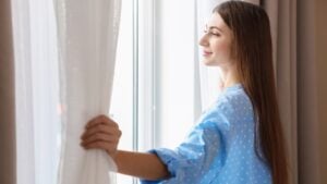 Smiling woman opening window curtains at home