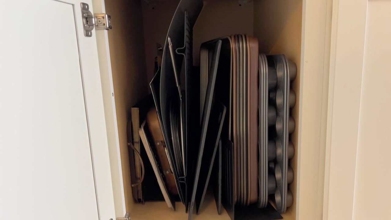 A neatly arranged kitchen cabinet with vertical storage for baking sheets, muffin tins, cooling racks, and baking pans.