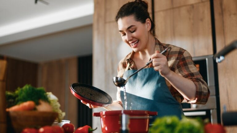 A joyful woman enjoying her time cooking in a modern kitchen. Surrounded by fresh vegetables, she uses a red pot and ladle to prepare a meal, exemplifying the art of home cooking and culinary creativity.