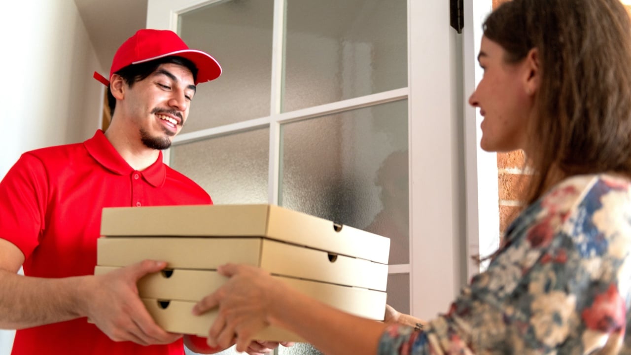 A delivery person in a red uniform hands pizza boxes to a customer at the door. The setting is a bright entryway with a glass-paneled door.