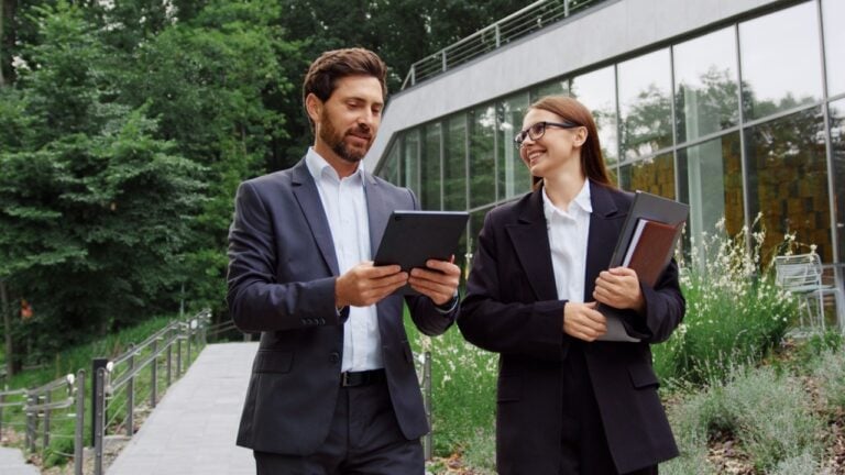 Confident business partners, a man and a woman in formal suits, are walking and talking near a modern office building, with the man using a laptop and the woman holding a tablet as they discuss ideas
