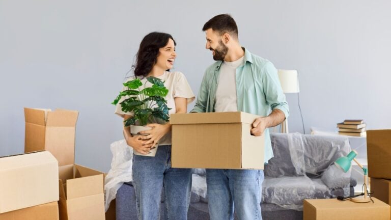 Couple moving with boxes in new apartment. Smiling happy family looks up together, planning their home. Unpacking boxes, sofa and houseplant. Relocating, real estate and mortgage concept.