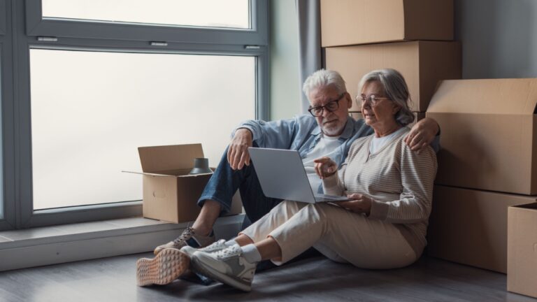 Elderly couple sits on the floor surrounded by cardboard boxes, using a laptop to plan moving details, share memories and make decisions together during a calm, intimate home transition.