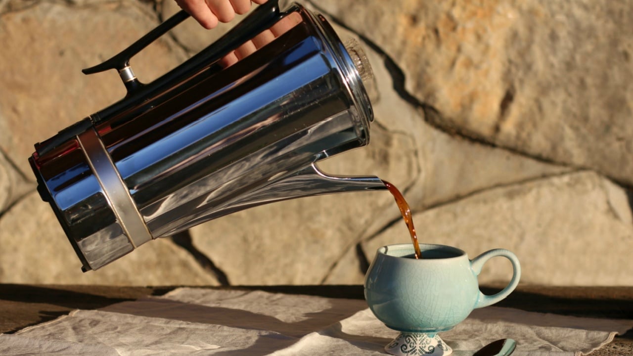 Dark, Black Coffee Being Poured From Vintage Metal Coffee Percolator Into Blue Ceramic Cup On Wooden Table With All Natural Cotton Linen Tea Towel And Spoon And Large Stone Background