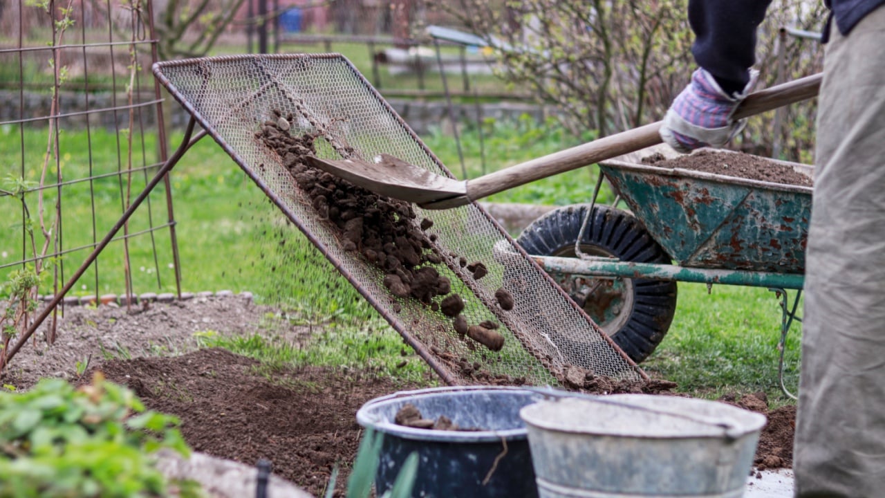 Adult man works on garden. Man shovel soil through homemade screen for sorting on stones and clay. Homemade device for sifting clay. Shovel, wheel. Landscaping work. Hard working man. Manual work.