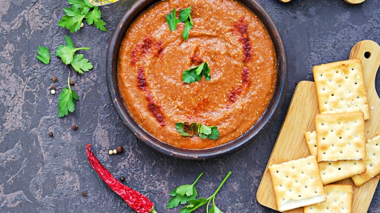 Appetizer hummus red lentil and red pepper in a clay bowl on a dark concrete background. Served with salted crackers and chopped vegetables.