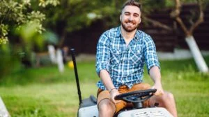 lawncare concept - handsome young man trimming grass in garden using tractor