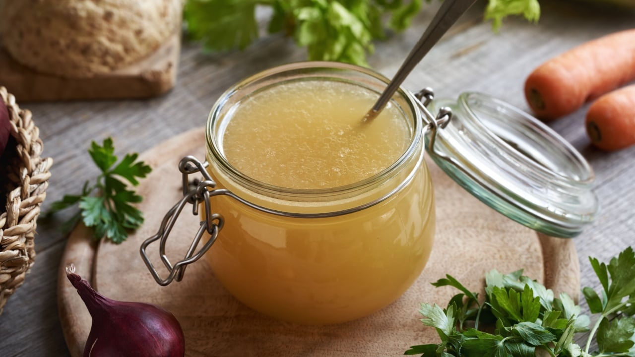 Chicken bone broth or soup in a glass jar on a table, with fresh vegetables and herbs