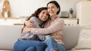 Two beautiful women, older mother and young millennial daughter embracing relaxing seated on cozy sofa in modern living room, enjoy life moments, feeling unconditional love, appreciate time together