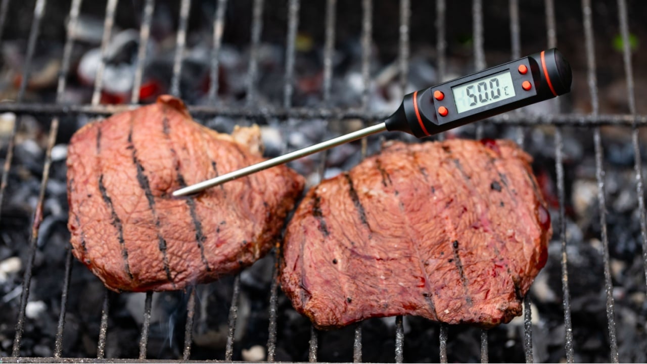 Close-up shot of digital meat thermometer inside grilled steak on barbecue showing 50 degrees Celsius for rare doneness of beef