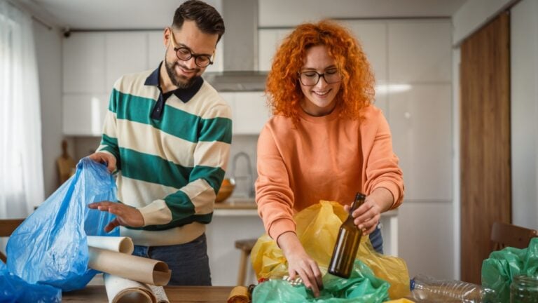 wife and husband sit on wooden desk recycle at home sorting waste plastic paper and glass to green, yellow and blue bags sustainable living concept