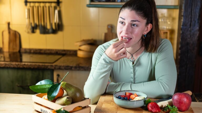 Woman leaning on kitchen table, eating fruit from a bowl while surrounded by fresh fruit. Warm and natural scene with soft light.