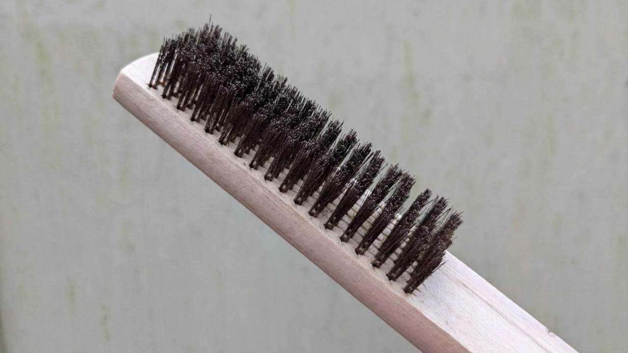 A close-up shows a wire brush with stiff metal bristles set in a light wooden handle against a blurred, textured background.