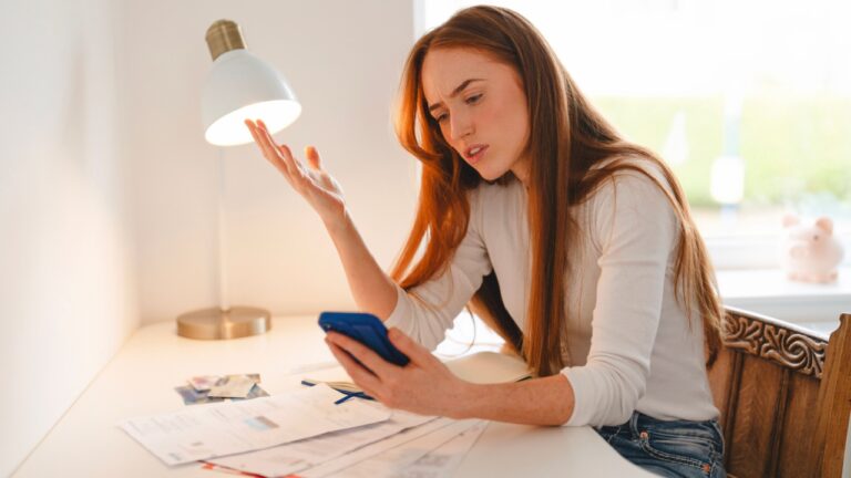 Woman looking frustrated while using a smartphone at a table with bills and paperwork during daylight in a cozy indoor setting