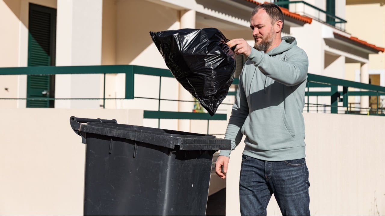 Individual is placing a black trash bag into a bin on a clear day in a residential area