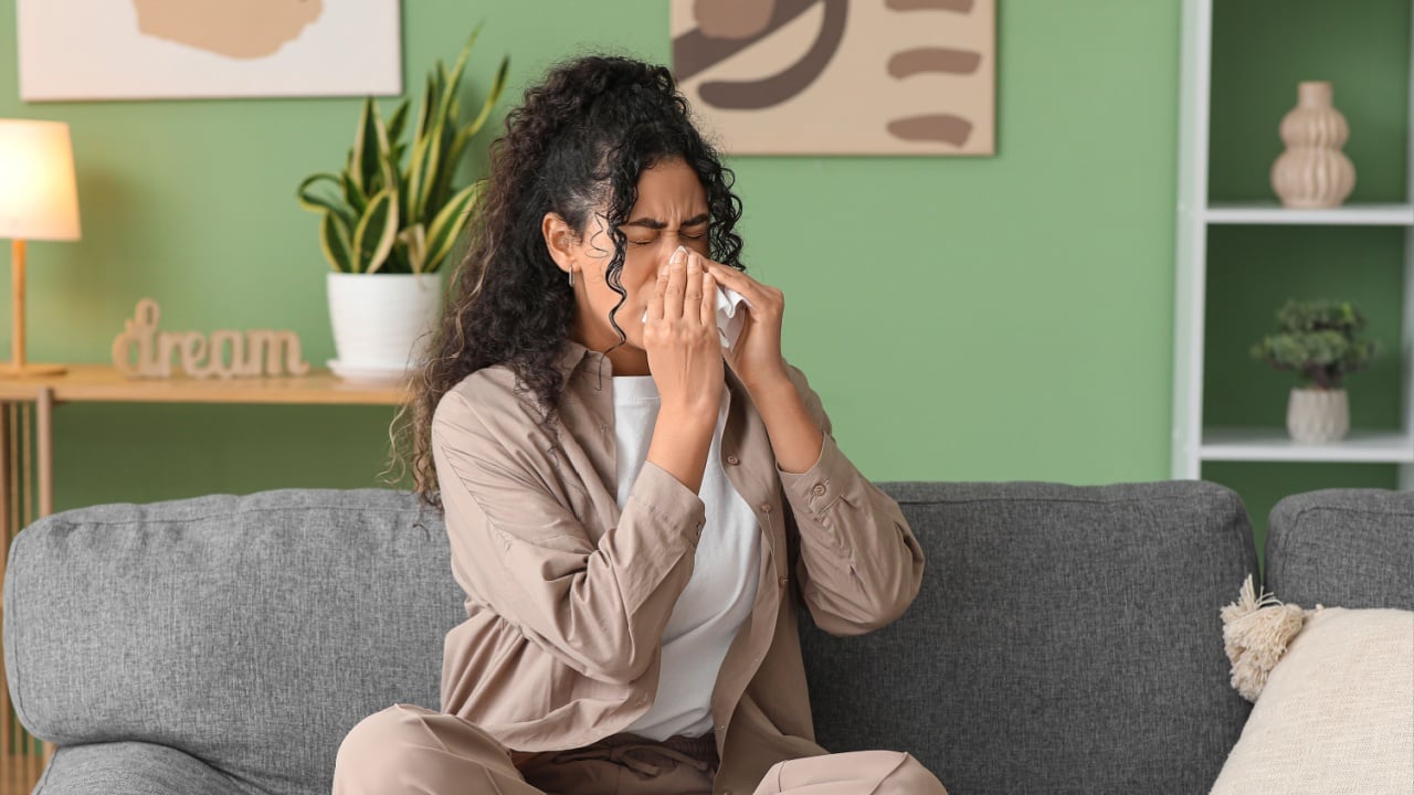 Young African-American woman sneezing in handkerchief and sitting on sofa at home. Allergy concept