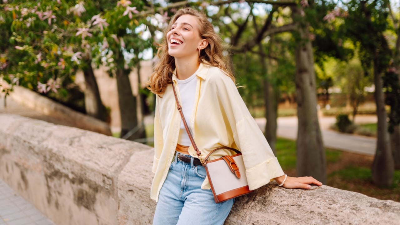A beautiful woman in stylish clothes strolls along sunny city streets, admiring the blossoming trees. A young woman strolls through a park in the sun's rays. Concept of a walk, spring, and beauty.