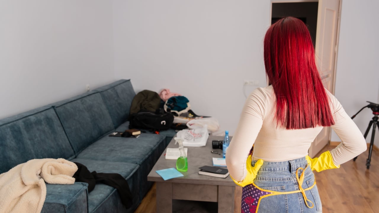 Young woman looking frustrated and angry while standing in messy untidy bedroom full of scattered clothes and objects, expressing annoyance about household disorder.