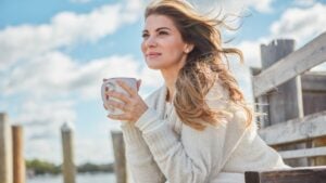 Thinking, bench and woman with coffee cup on pier, holiday reflection and positive mindset for peace. Calm, perspective and person with smile for vacation, tea drink and outdoor for daydreaming