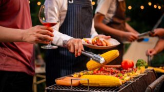 An Asian woman with friends grilling BBQ skewers, corn, and sausages outdoors. They toast with wine amid glowing string lights, creating a joyful and warm evening barbecue atmosphere