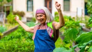 Happy senior woman dancing with arms raised in her home garden, celebrating an active and joyful retirement while cultivating fresh vegetables outdoors