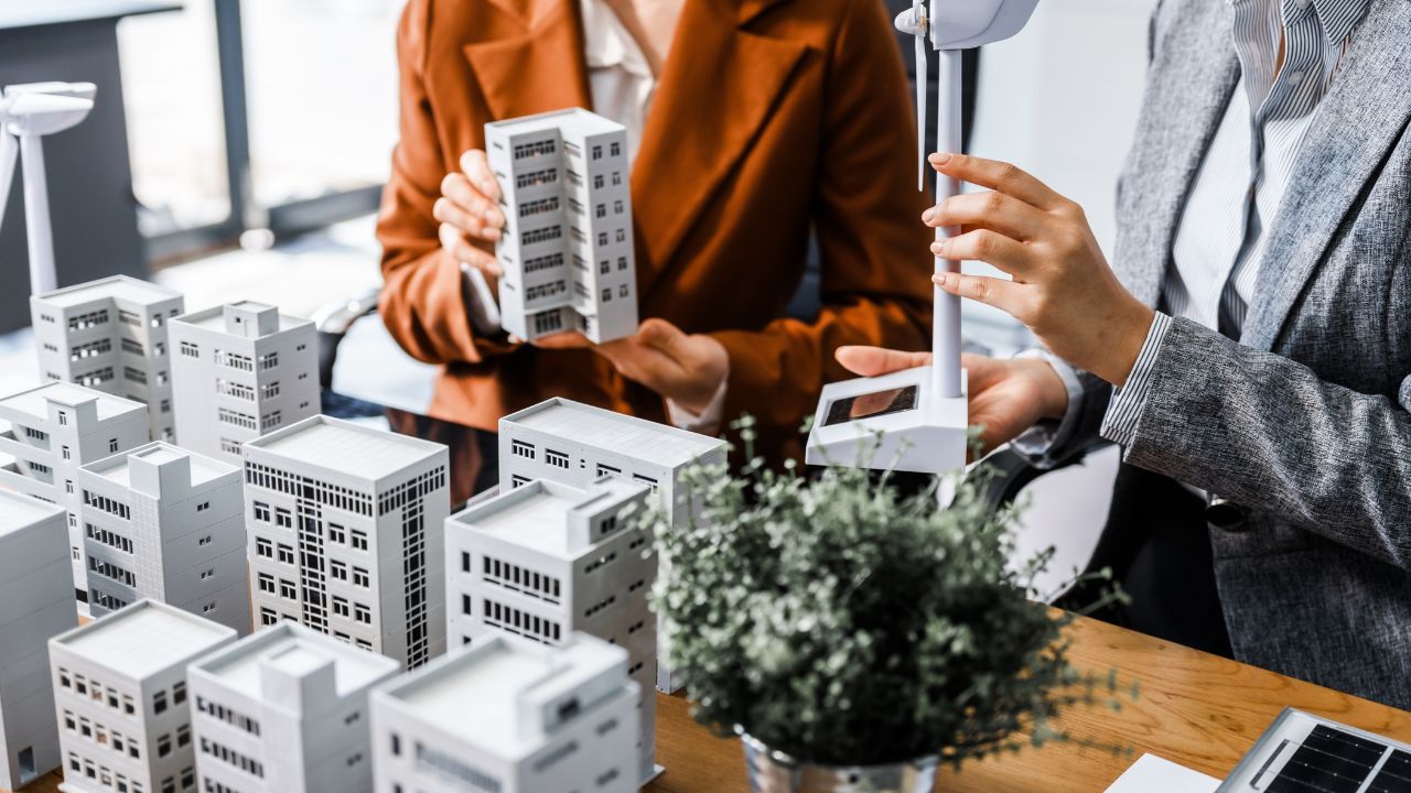 Two businesswomen sit at a desk in a meeting, discussing property development while building a model of city tower. real estate investments, zoning laws, sustainable energy solutions. net zero