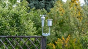 A small weather station with anemometer and rain gauge stands above a wooden fence. Dense shrubs and trees create a lush garden monitoring scene.
