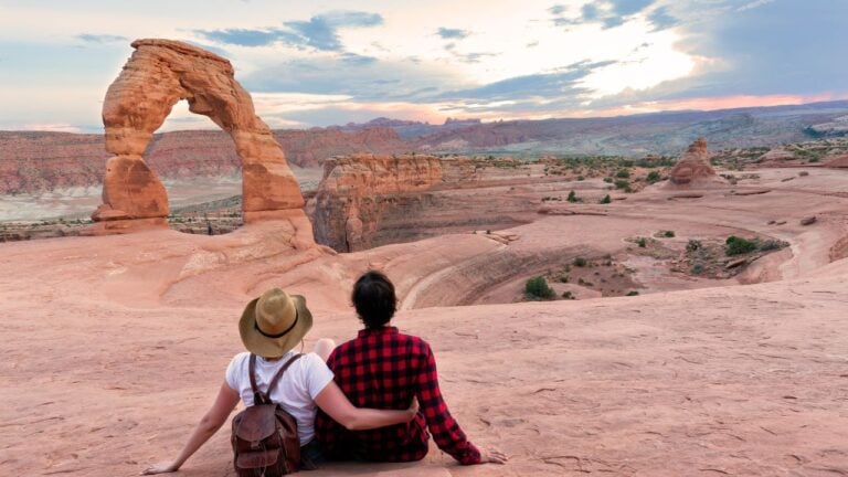 Couple on vacation hiking trip. Man and woman sitting on top of the mountain looking at beautiful view. Delicate Arch, Moab, Utah, Arches National Park.