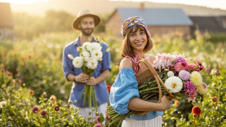 Portrait of cheerful man and woman stand as farmers with buckets full of freshly picked up dahlias while working at flower farm outdoors on sunset